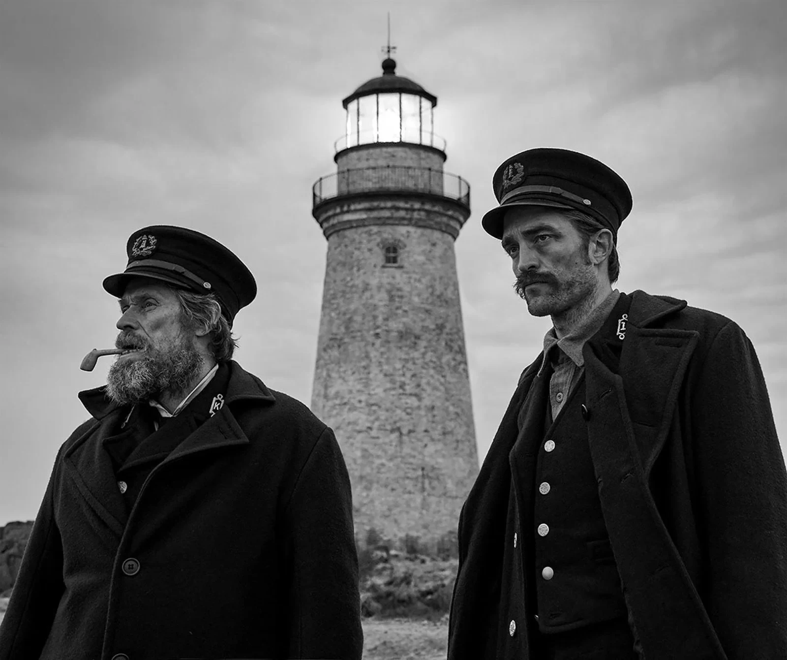 Willem Dafoe y Robert Pattinson con gorras de marinero frente a un faro, en blanco y negro
