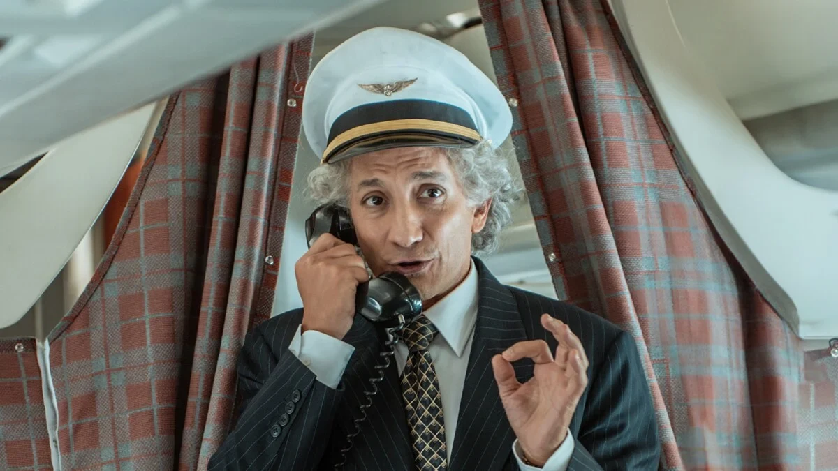 Juan Minujín, con gorra de piloto, habla por un teléfono de cable a bordo de un avión