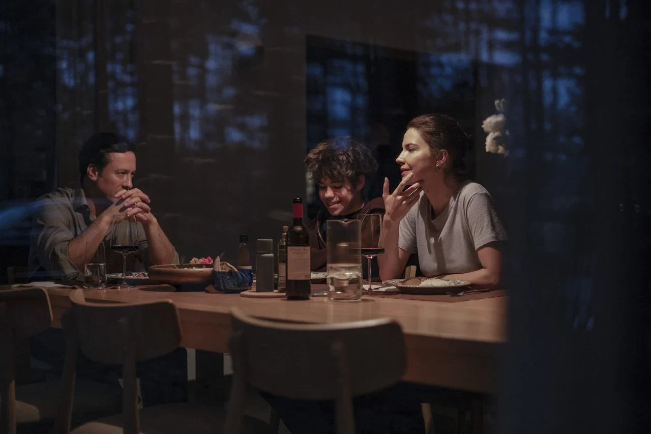 Tres personas conversan en una mesa de un restaurante, vistas a través de un vidrio