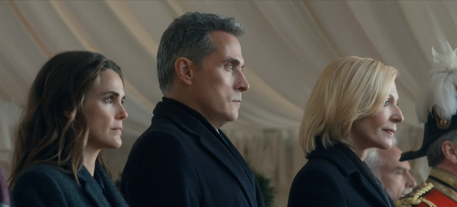 Keri Russell, Rufus Sewell y Allison Janney de perfil en una ceremonia al aire libre, en La diplomática