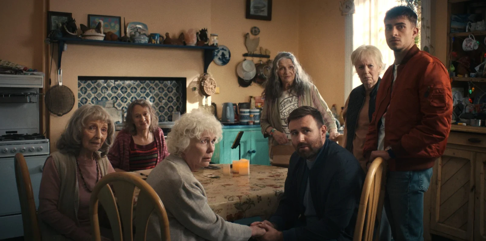 James McArdle toma de la mano a Fionnula Flanagan en una mesa de cocina, rodeados por otras señoras, en Cuatro madres