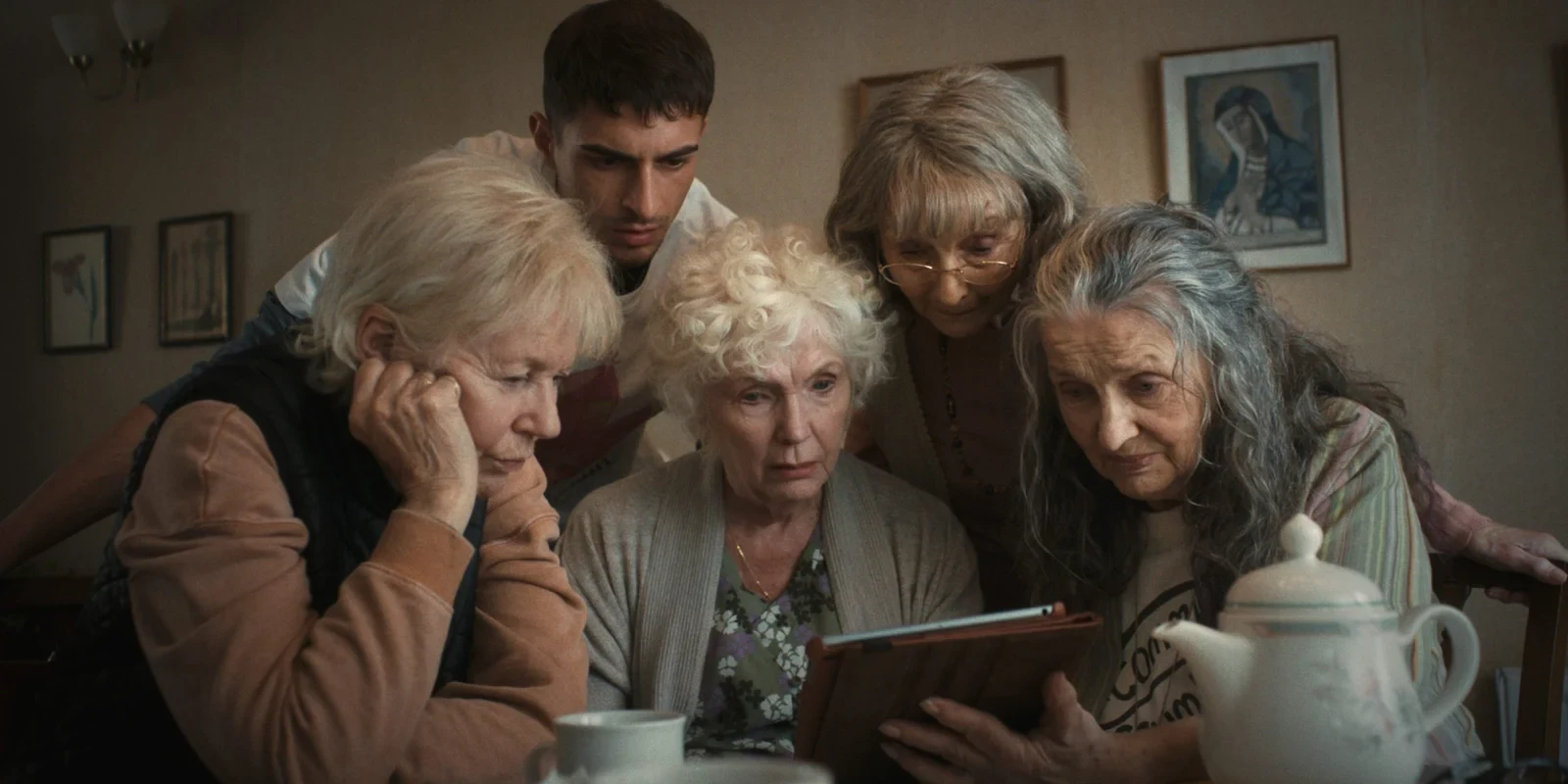 Cuatro señoras mayores y un joven miran juntos una tablet en un living, en Cuatro madres