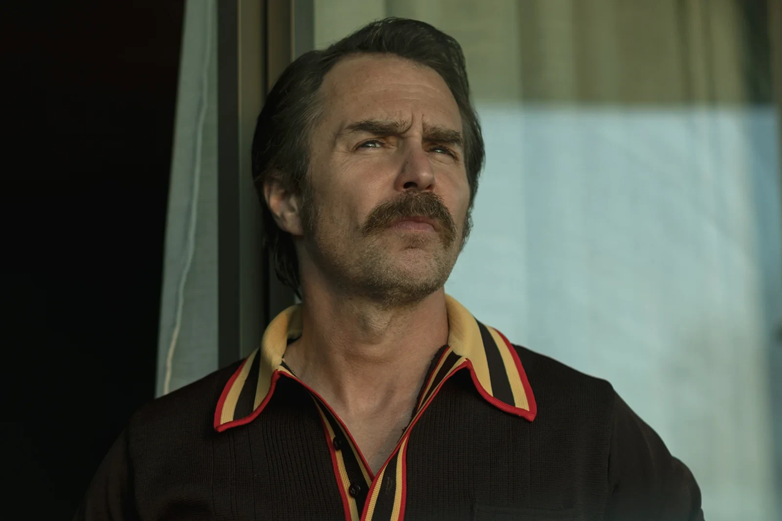 Sam Rockwell con bigote y camisa a rayas junto a una ventana