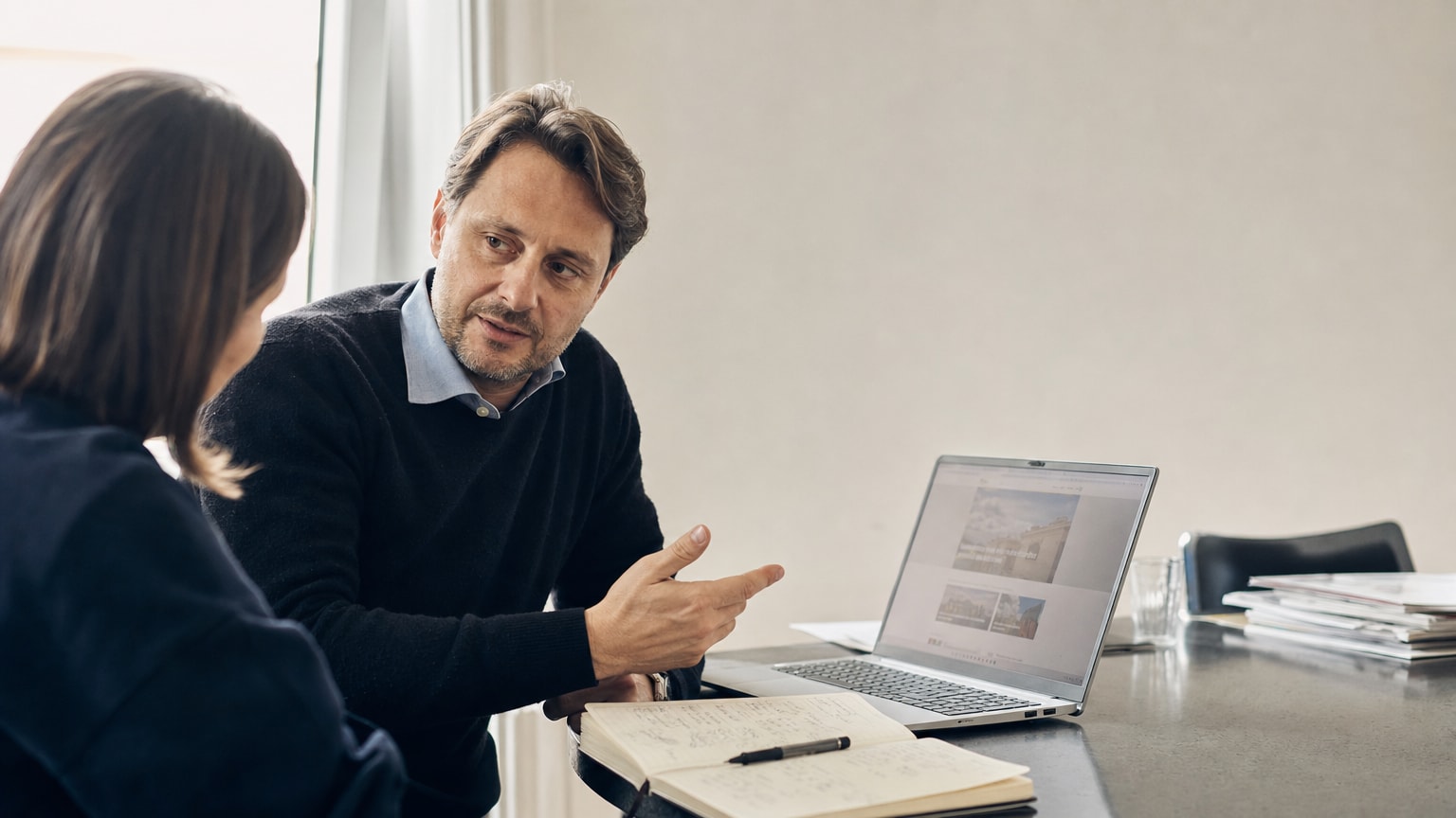 Ein aufgeräumter Schreibtisch mit einem Laptop, auf dessen Bildschirm eine moderne, klar strukturierte Website zu sehen ist. Daneben liegt ein Notizbuch mit handschriftlichen Textskizzen. Warmes, natürliches Licht, professionelle Atmosphäre ohne Werbe-Ästhetik.