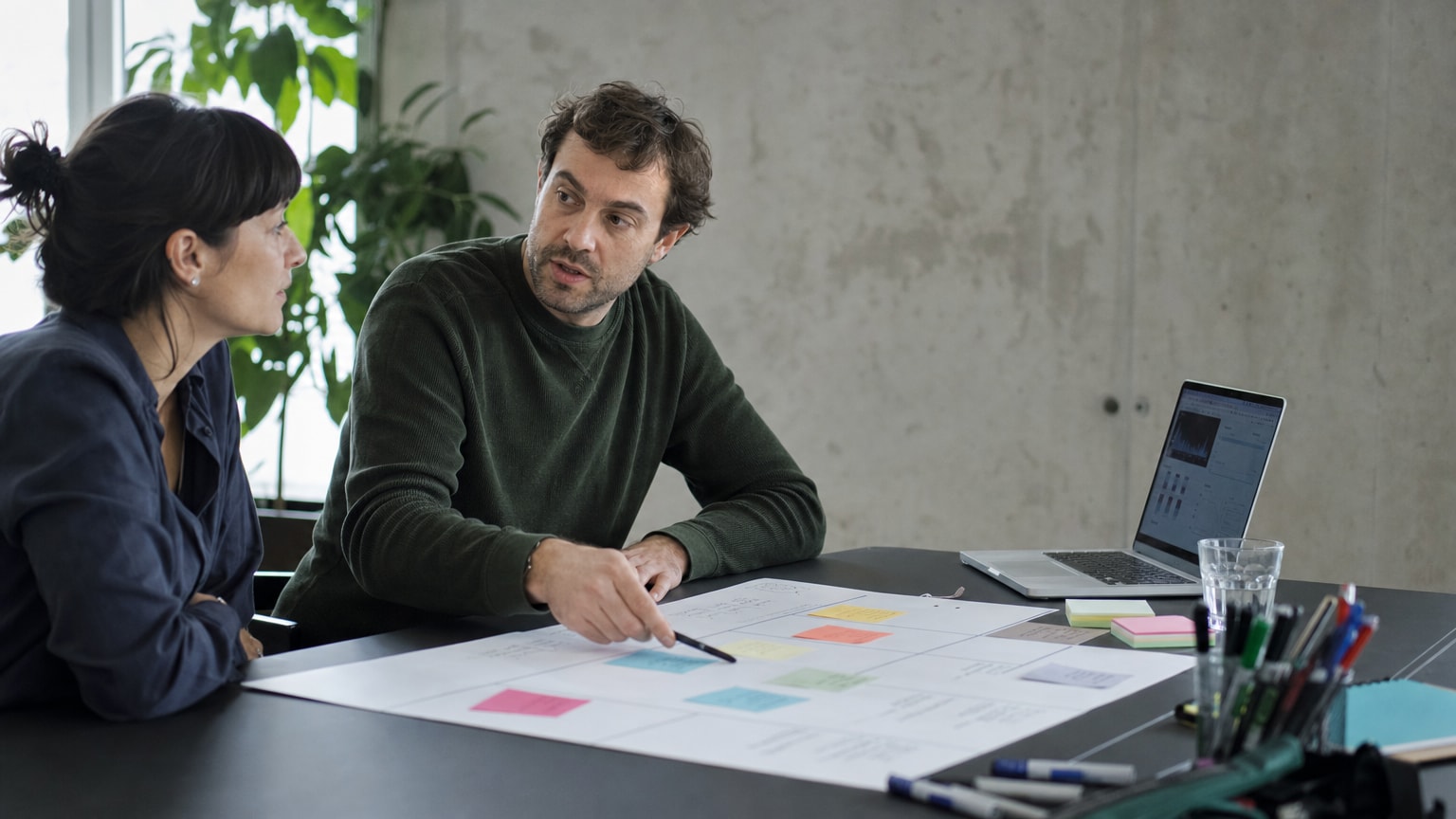 Ein aufgeräumter Schreibtisch mit einem einzelnen A3-Blatt, auf dem ein strukturiertes Canvas-Template mit farbigen Post-its und Stiften liegt. Im Hintergrund ein Laptop mit Analytics-Dashboard, helle, professionelle Atmosphäre.