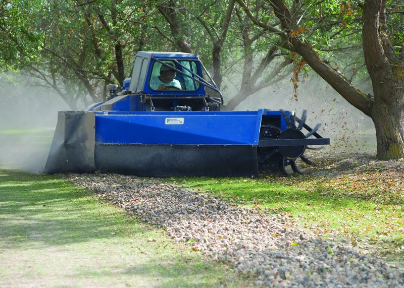 A blue self-propelled sweeper machine gathers almonds into a windrow in a California orchard, throwing up dust as the operator drives between the tree rows
