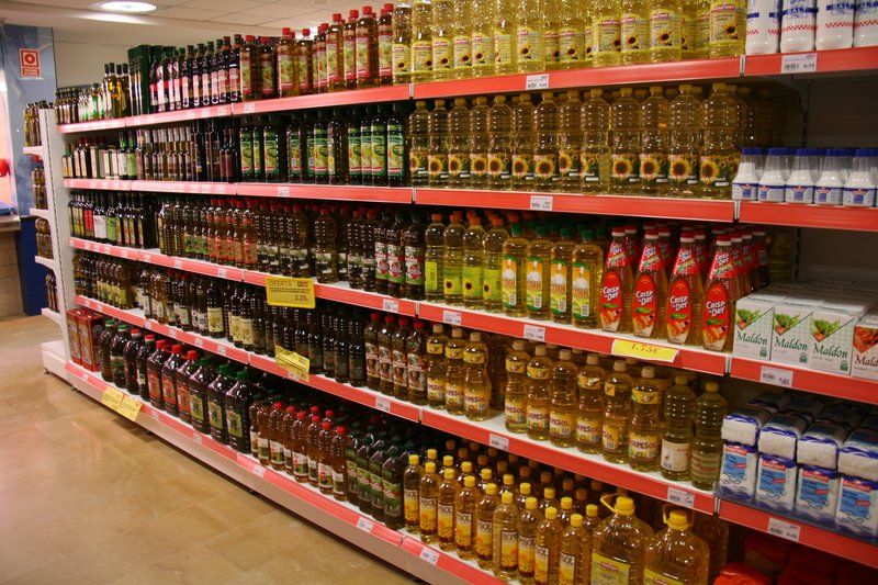 Supermarket shelves in Spain stacked with bottles of olive oil, including Carbonell labels, alongside sunflower oil and table salt