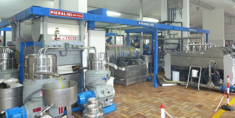 Stainless steel vessels, pumps and a continuous extraction line inside a modern olive oil mill in San Miniato, Tuscany, with pale green oil running into an open tank