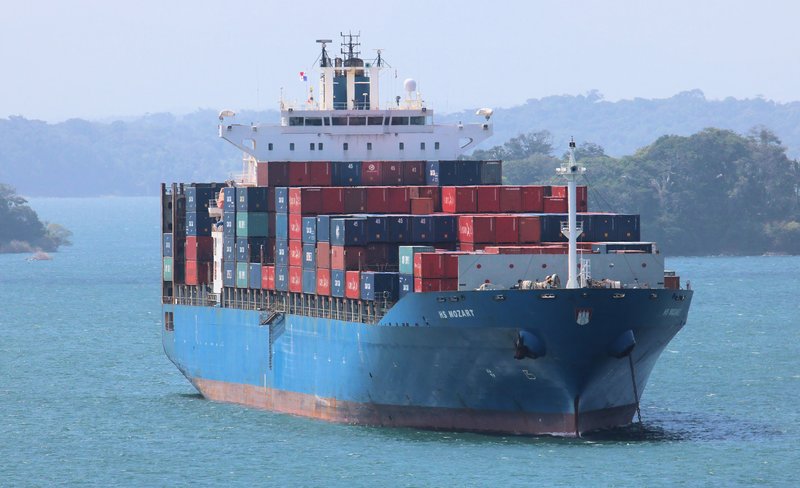 A blue-hulled container ship stacked high with containers under way on the open water of Gatun Lake, forested hills on both banks