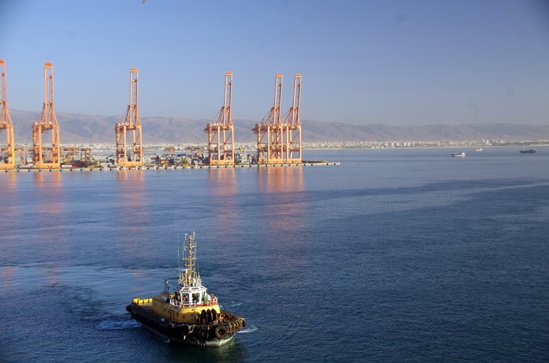 Gantry cranes and rows of stacked containers along the quay at the port of Salalah, Oman, with a tug under way in the foreground