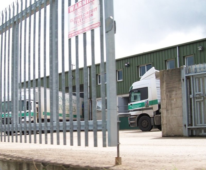 Lorries in Glens of Antrim Potatoes livery parked at the company's green-clad depot, seen through a steel palisade fence with a CCTV warning sign
