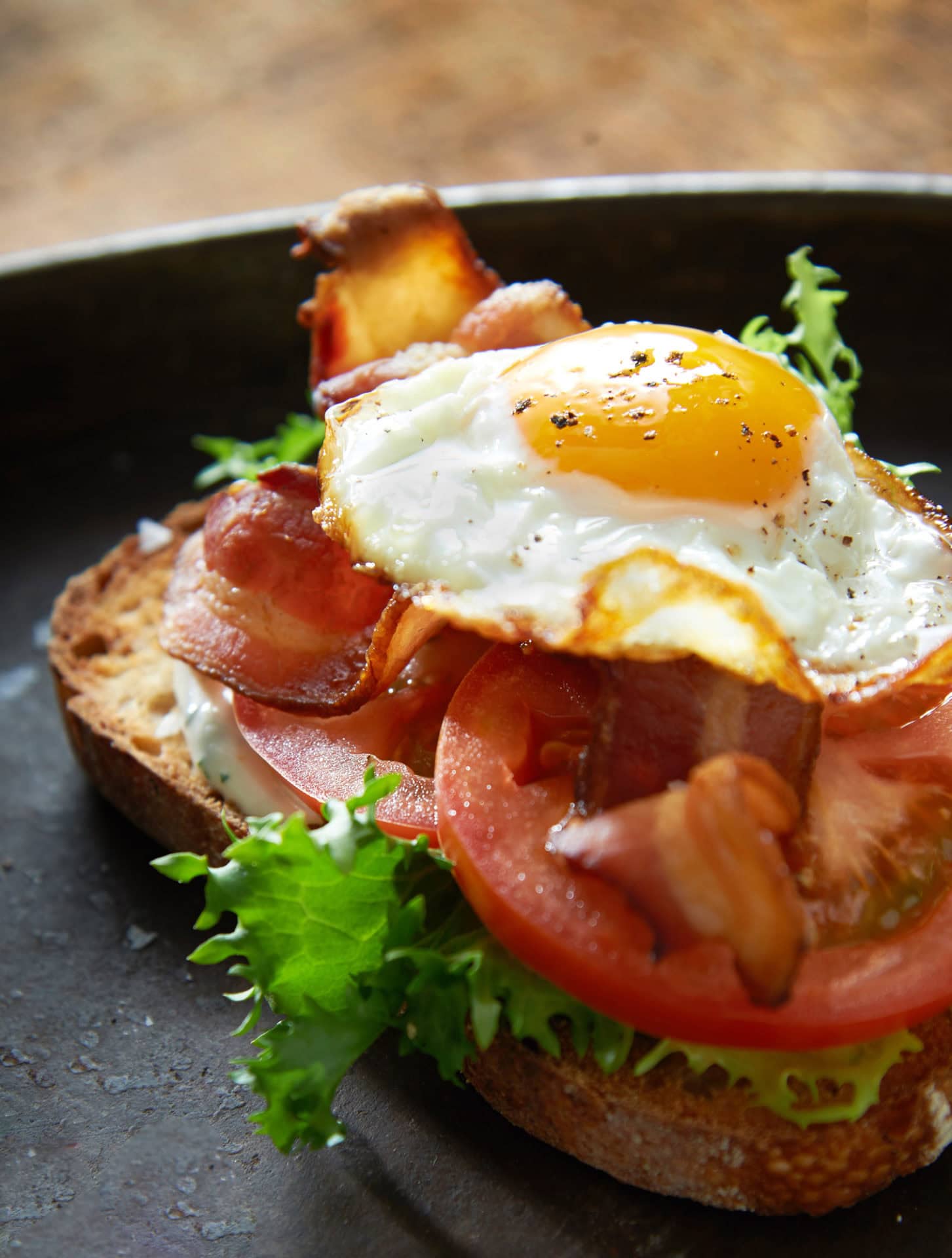 open faced sandwich with toasted bread, bacon, tomato, lettuce and fried egg on a black plate