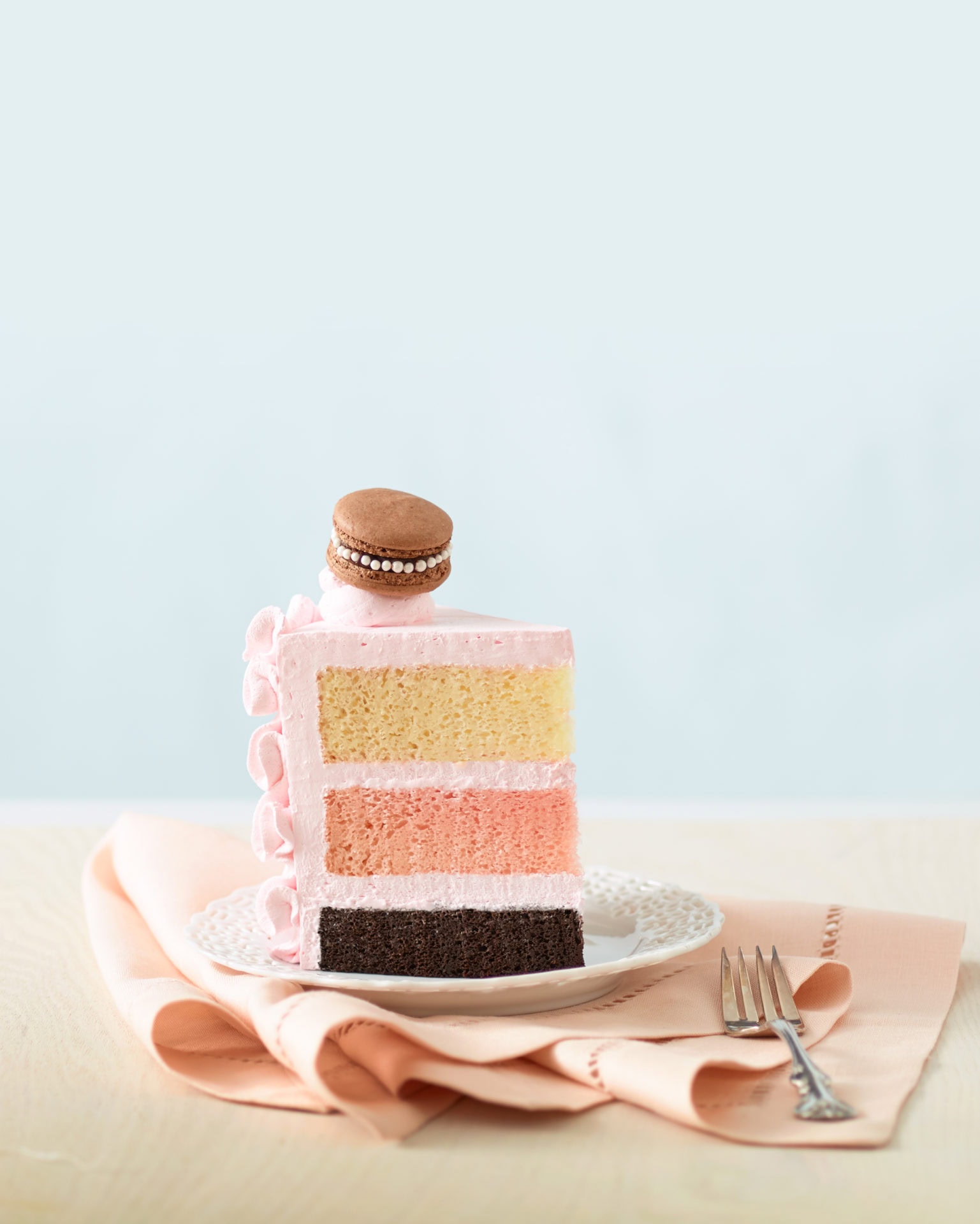 cake slice on a table and plate.  cake has light pink frosting and 3 layers of chocolate, strawberry, and vanilla on top.  decorated with a brown macron. 