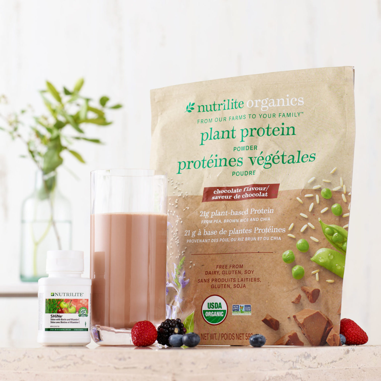 bag of plant protein smoothie mix on table with berries and glass of smoothie.  Also a bottle of supplements on the table. 