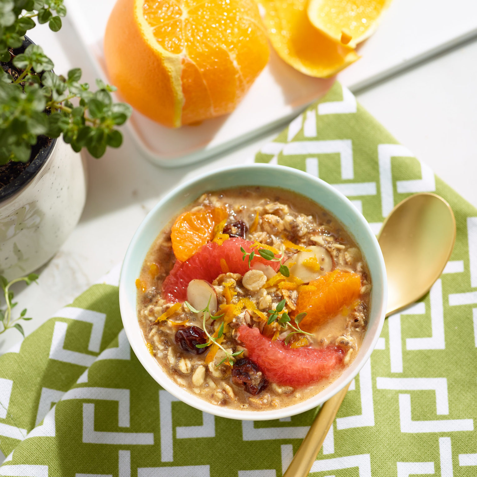 bowl of oatmeal with berries, seeds, grapefruit and oranges on top.  Bowl is on a table with green and white patterned napkin, gold spoon, cut oranges and a plant in the top left corner