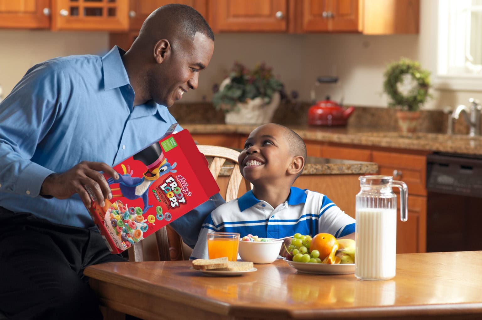 dad with son at kitchen table. dad is pouring a bowl of fruit loops cereal for son. 