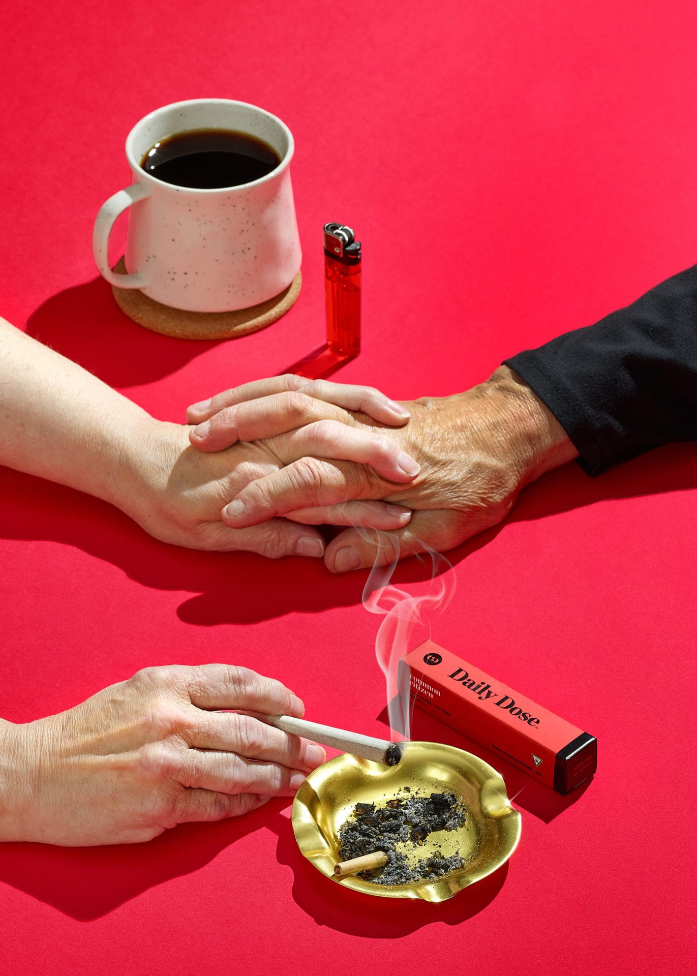 hands of woman holding lit cannabis joint over an ashtray while also holding a mans hand - on red table.  also on table is a cup of coffee, red lighter, and red packaging. 