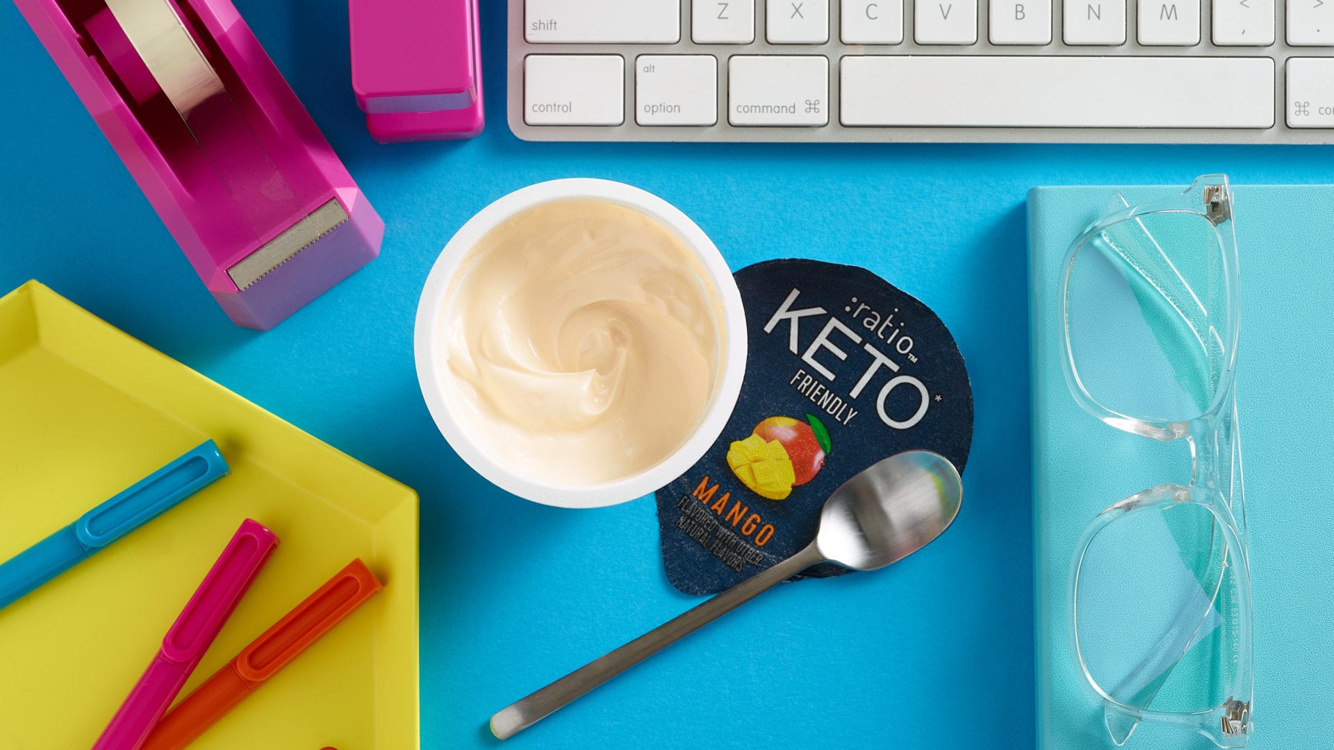 keto yogurt cup on blue desk with spoon, glasses, colorful pens, pink tape dispenser and part of a white keyboard