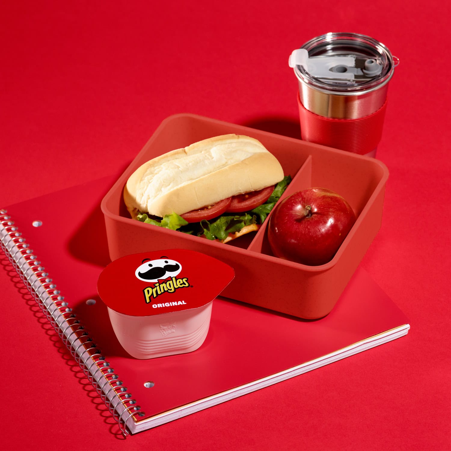 boxed lunch with water cup and red notebook and small package of red covered pringles