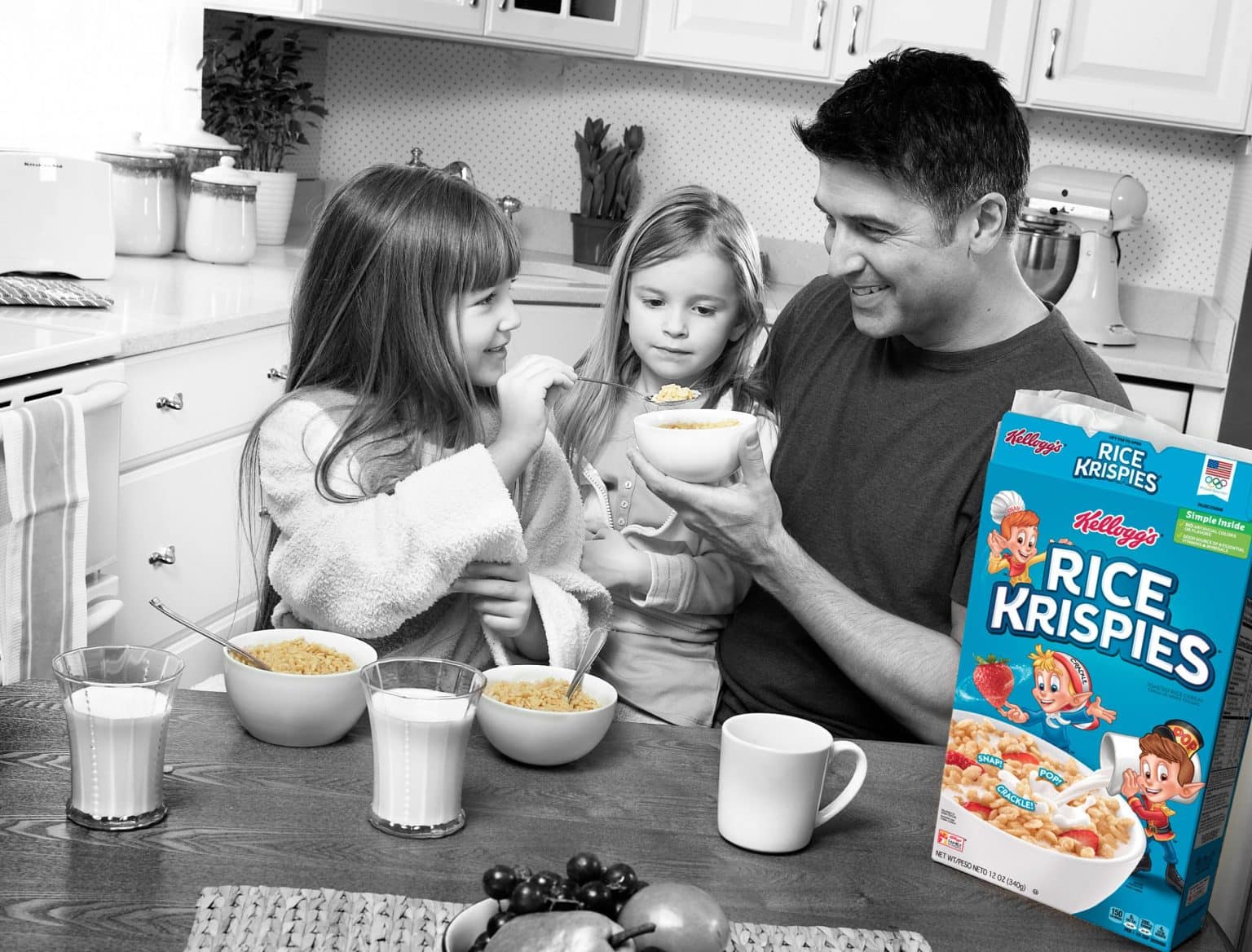 dad with 2 daughters eating bowls of rice krispies in kitchen.  Everything is in black and white except the cereal and box on table. 