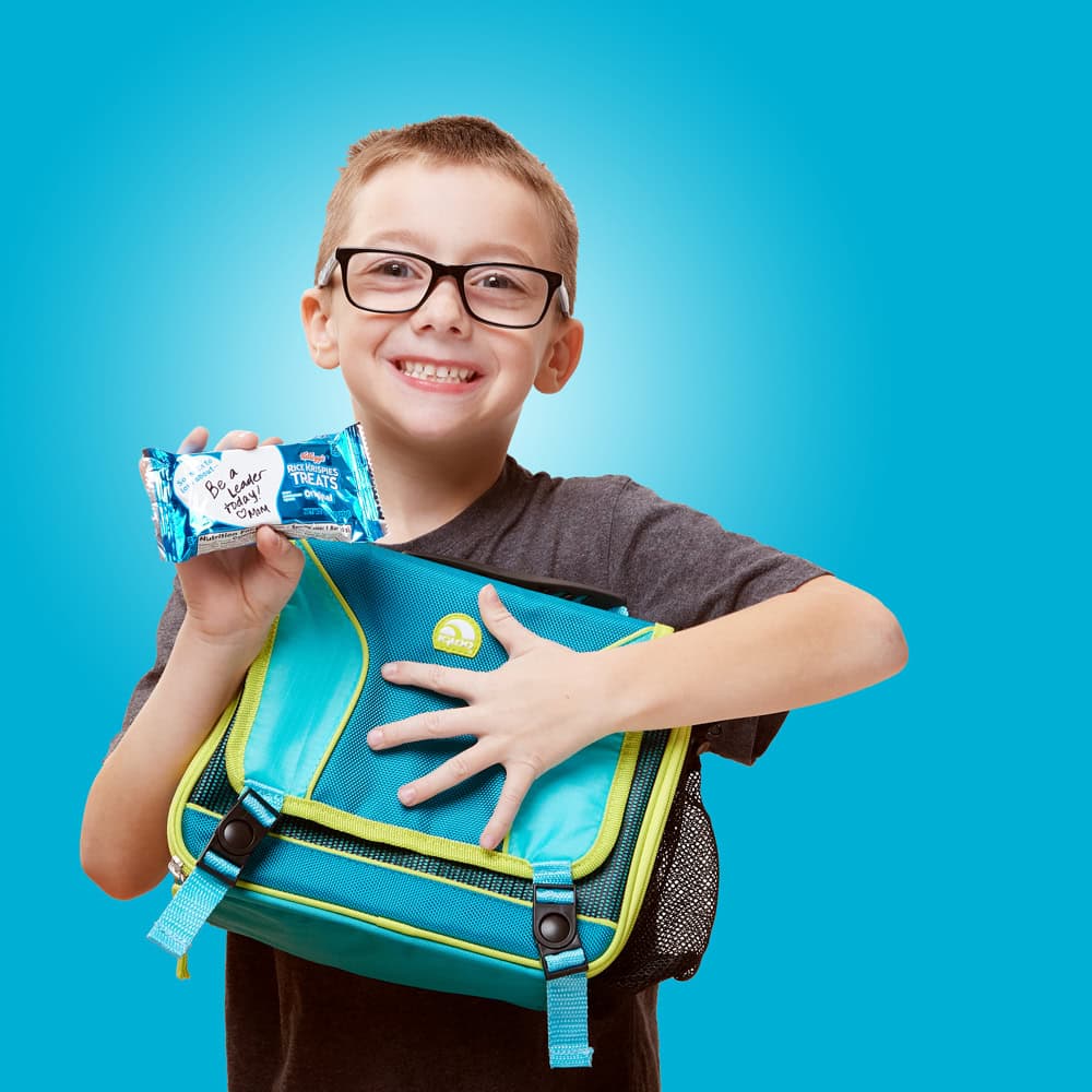 photo of boy holding backpack and rice krispies treat in package with blue background