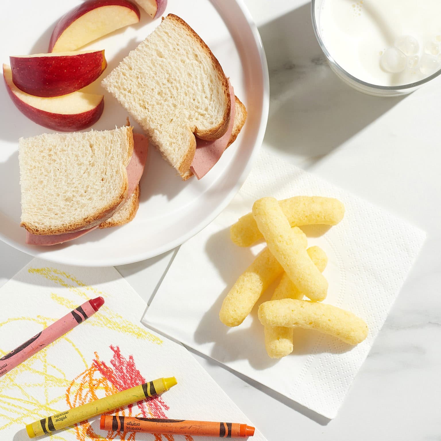 cheese puffs on napkin on table with a plate with sandwich and cut apples, crayons and paper