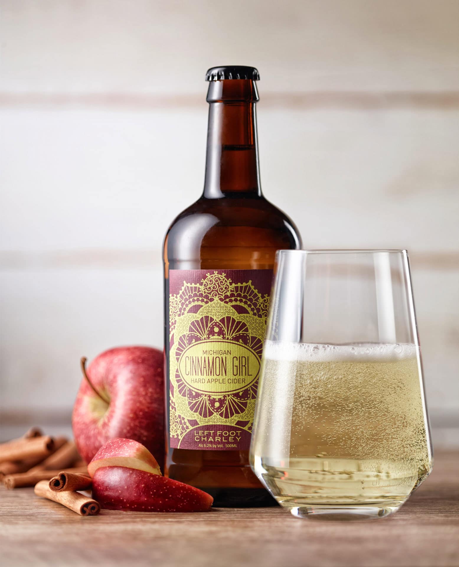 bottle of cider on table with glass of cider  next to it along with apples and cinnamon sticks