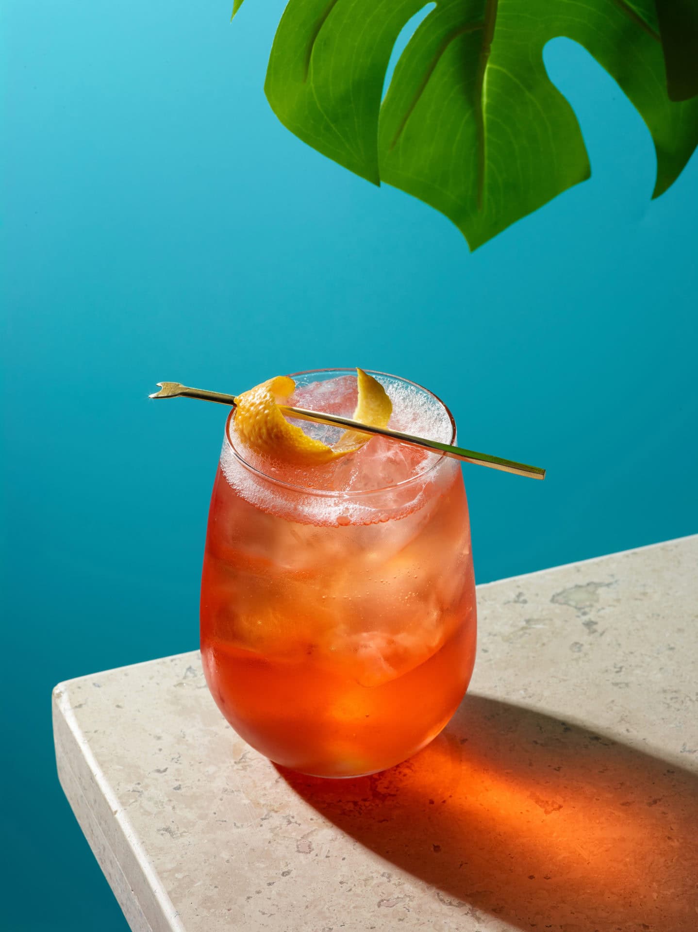 aperol spritz drink with lemon wedge on a table, blue background and a plant leaf in top right corner 