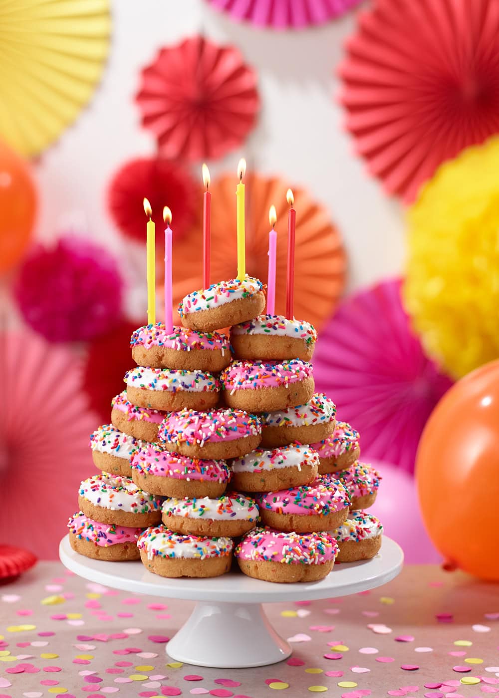 stack of donuts on a cake plate, white glaze with sprinkles.  topped with colorful candles.  Background of colorful paper decorations and balloons in a birthday party setting. 