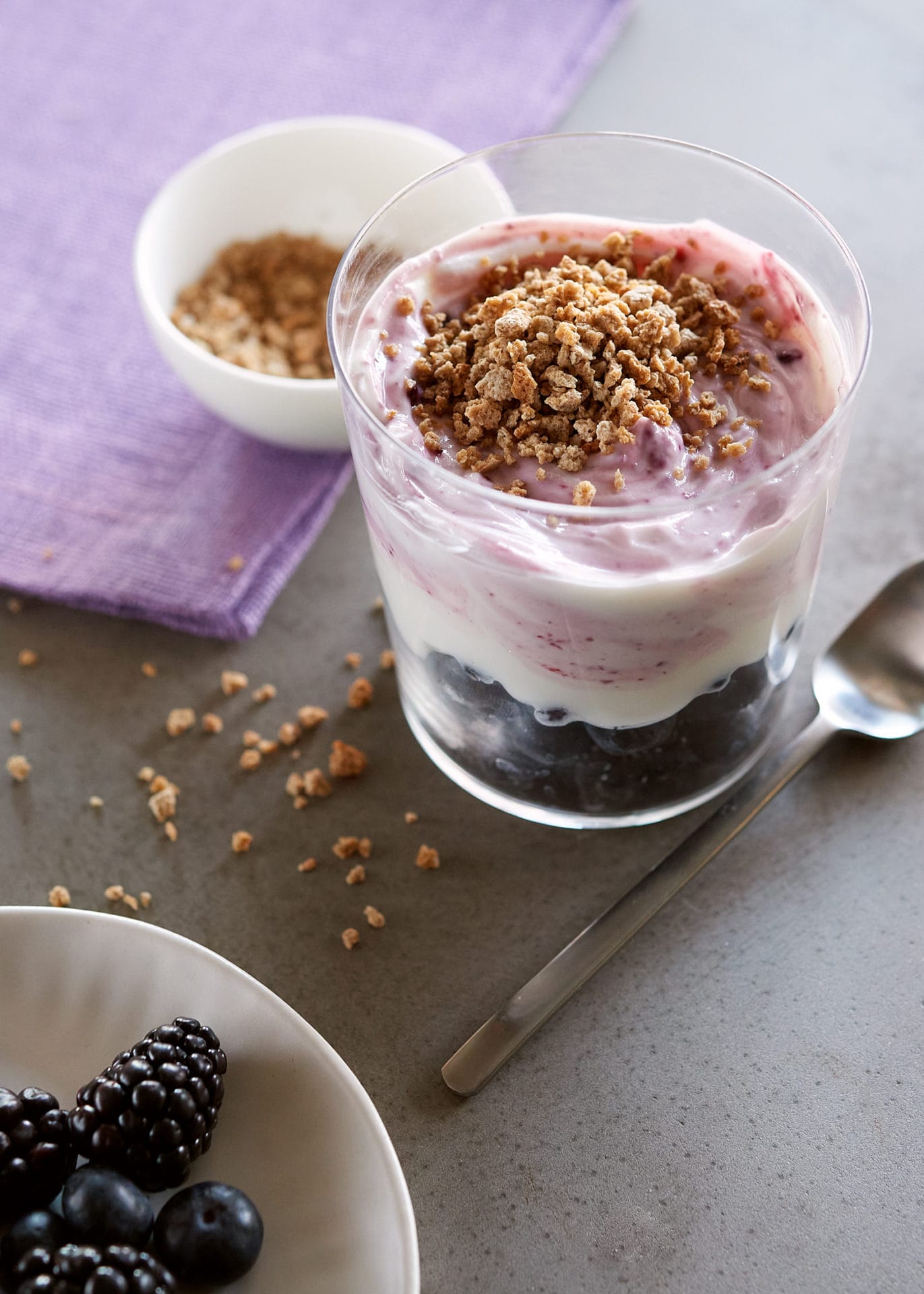 parfait with berries, yogurt and granola on a table with spoon, dish of berries etc. 