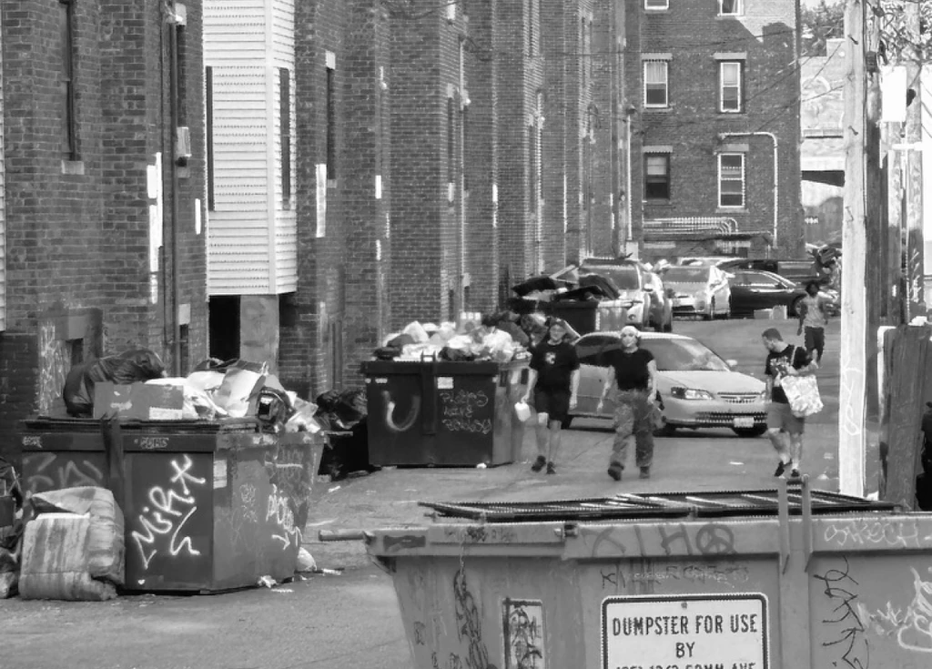 An Allston alley of overflowing dumpsters and discarded mattresses as students pass