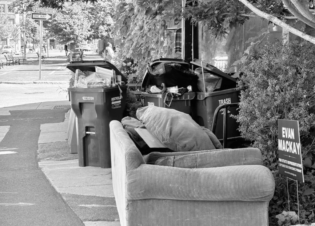 A corduroy sofa left on the sidewalk beside overflowing trash and recycling bins
