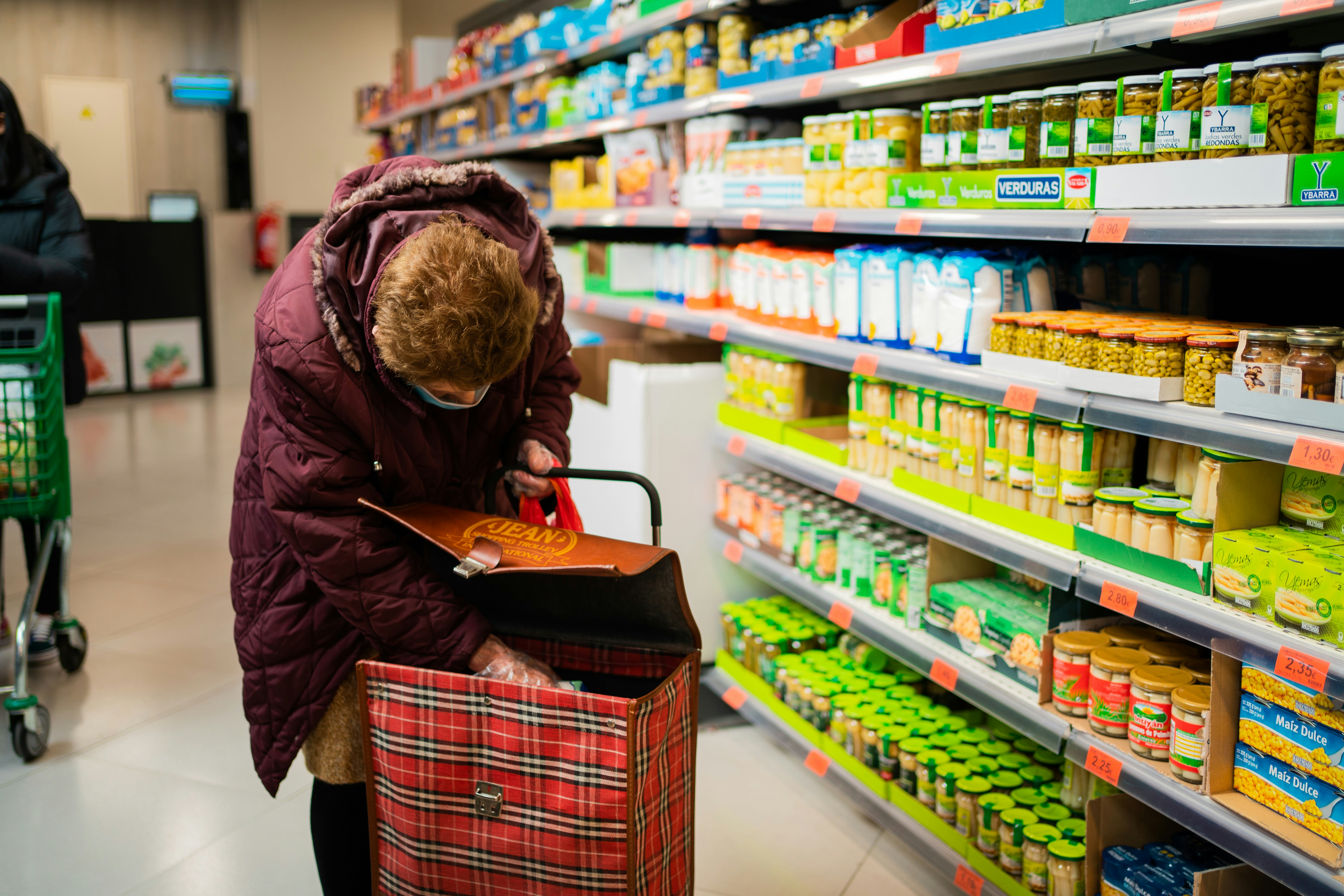 Elderly woman loads her personal wheel cart with groceries in an aisle.