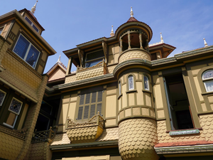 An ornate turret and textured exterior walls of the Winchester Mystery House showcase intricate Victorian architecture under a clear blue sky. Stained glass windows and decorative trim add to the surreal beauty, while a sign reading "Door to Nowhere" marks a famously strange and haunting feature of the mansion. The whimsical design hints at the home's mysterious history and eccentric construction.