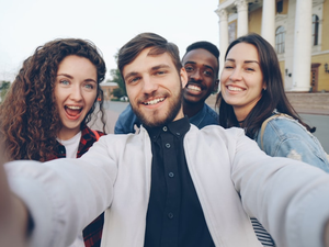 Point of view shot of multiracial group of tourists taking selfie in city center holding gadget camera and posing together with hand gestures expressing positive emotions.
