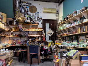 Traditional Chinese Tea Shop With Shelves Of Ceramic Teapots And Teacups - A small Chinese tea shop with shelves filled with ceramic teapots and teacups. A young girl sits in a chair by a table. The shop is cluttered with boxes and other items.