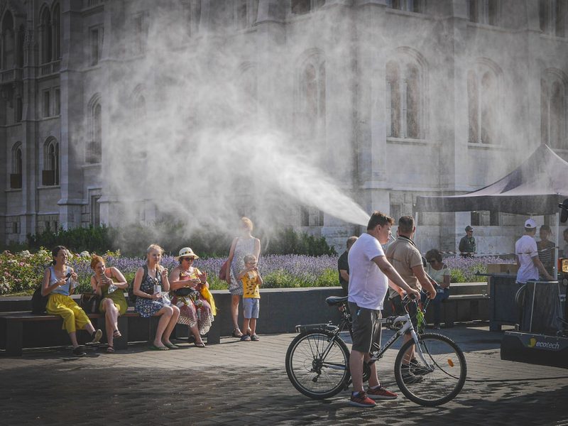 Street scene on a hot summer day in Budapest with a cyclist passing through a cooling mist, people sitting on benches and enjoying the shade. A humorous and refreshing moment of everyday life.