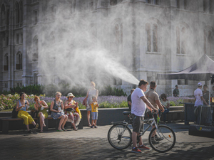 Street scene on a hot summer day in Budapest with a cyclist passing through a cooling mist, people sitting on benches and enjoying the shade. A humorous and refreshing moment of everyday life.
