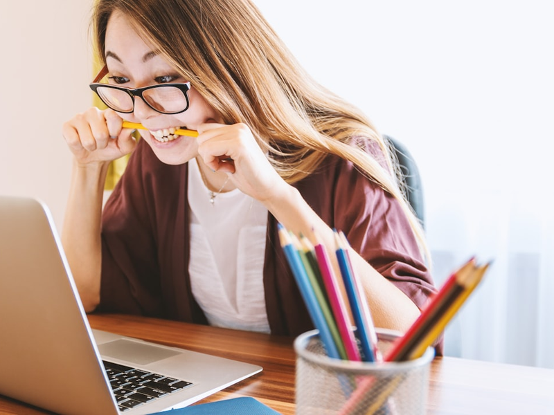 woman biting pencil while sitting on chair in front of computer during daytime