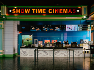 The lobby space of a cinema in Taichung, Taiwan features a vibrant "SHOW TIME CINEMAS" sign in bold, red letters overhead. Below, a snack bar or concessions counter awaits, where a staff member prepares snacks. Digital menus display a variety of food and drink options, including popcorn and soft drinks. Black stanchions and ropes clearly define the queueing area for patrons, creating an organized atmosphere.