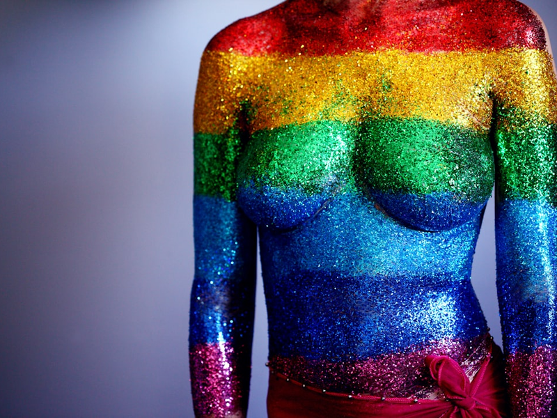 An 18 year old person covered in rainbow body glitter in support of the LGBTQ community and to celebrate gay pride 2018. We used a synthetic cosmetics brush to paint clear hair gel on their torso and then they laid down and I sprinkled the glitter on their body, one stripe at a time. We were able to touch up by dabbing more glitter on after they stood up. We used water-proof adhesive bandages to cover the sensitive areas on the chest and the belly-button.