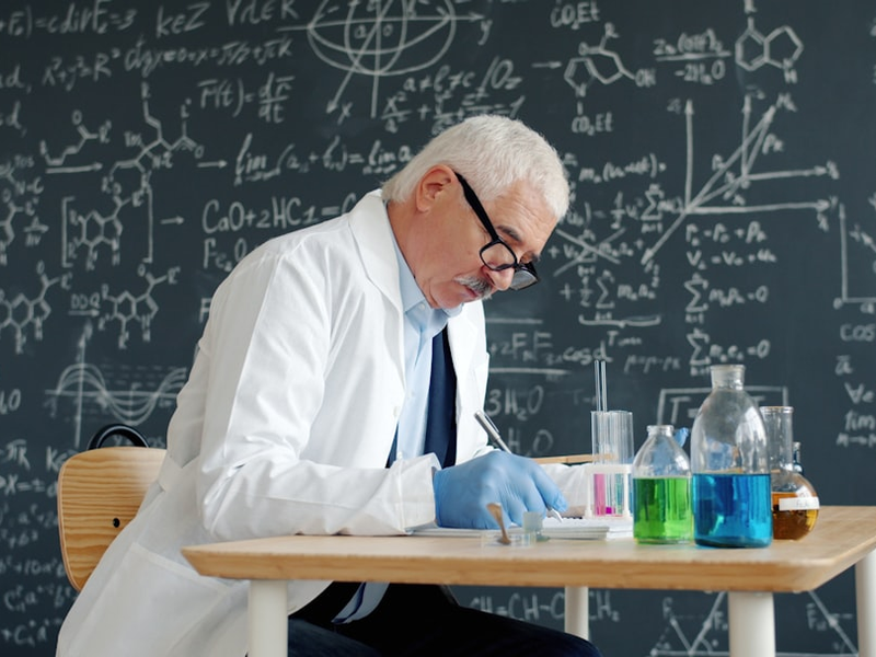 University professor wearing white gown, protective rubber gloves and glasses is writing in class at desk with chemical equipment glass tubes with colorful liquid.