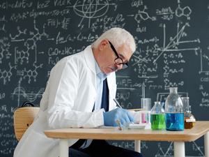 University professor wearing white gown, protective rubber gloves and glasses is writing in class at desk with chemical equipment glass tubes with colorful liquid.