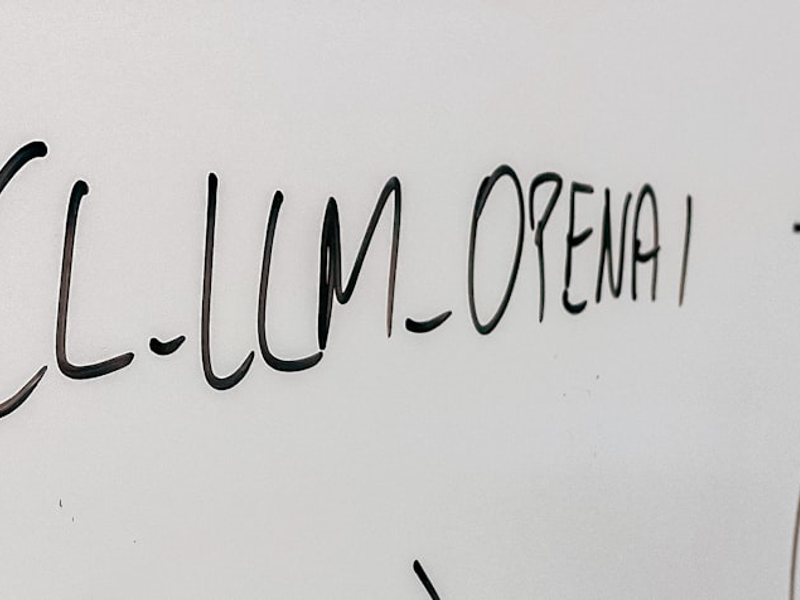 The image displays a white background with handwritten text. The primary text reads "LLM OPENAI" in black ink. There's also additional, somewhat blurred text in the background that is harder to decipher. The handwriting has a fluid and cursive style.