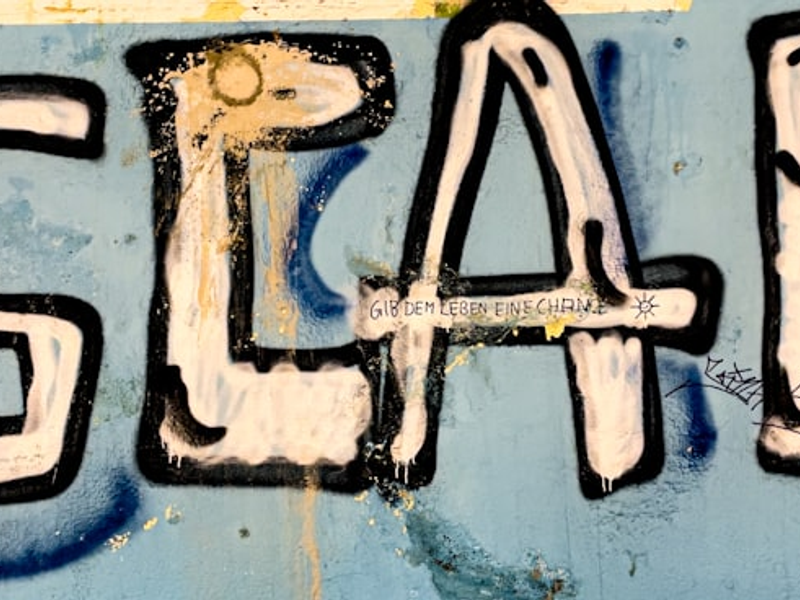 A graffiti piece on a weathered blue wall features the word "ESCAPE!" in large, bold, white and black letters. The paint appears to be dripping, and smaller handwritten text in German below it reads "Gib dem Leben eine Chance," which translates to "Give life a chance." The wall has visible signs of wear, including chipped paint and additional graffiti elements.