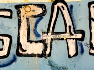 A graffiti piece on a weathered blue wall features the word "ESCAPE!" in large, bold, white and black letters. The paint appears to be dripping, and smaller handwritten text in German below it reads "Gib dem Leben eine Chance," which translates to "Give life a chance." The wall has visible signs of wear, including chipped paint and additional graffiti elements.