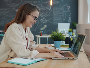Happy young lady is talking to friends online with laptop making internet video call waving hand sitting at desk in office. Communication and technology concept.