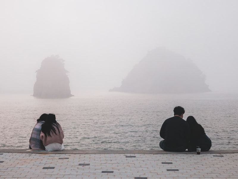 a man and a woman sitting on a stone walkway looking at a large rock in the water