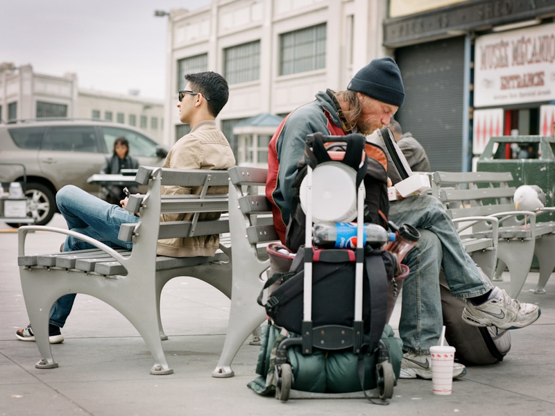 Hungry homeless man sits behind wealthy young man 