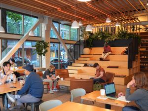 People work and read inside Powell’s City of Books, a Portland institution known for its cozy atmosphere and endless shelves. This café-like corner features tiered wood seating, indoor plants, and warm lighting, giving readers a place to relax or study. The storefront’s large windows let in daylight from the busy street outside.
